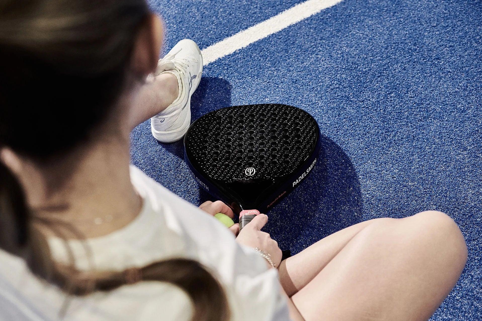 Woman sitting on blue padel court with black carbon padel racket and tennis ball