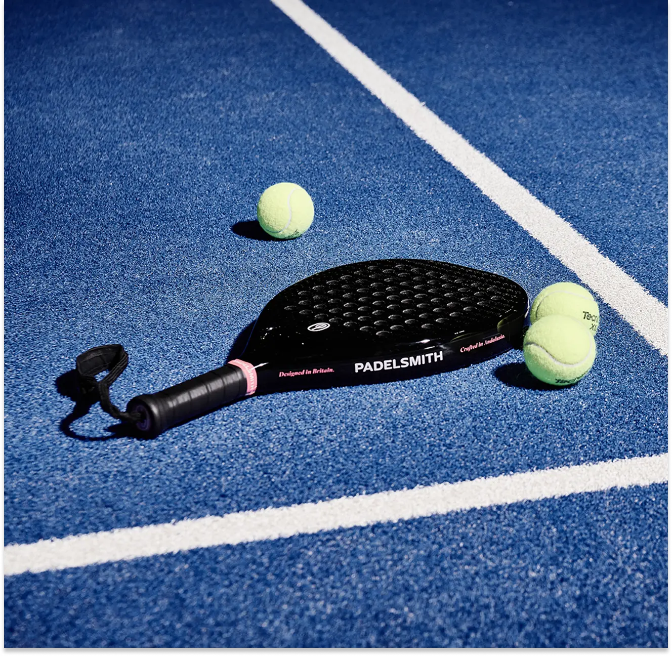 Padelsmith #1 padel racket lying on a blue court next to two padel balls, showing 'Designed in Britain, Crafted in Andalusia' branding
