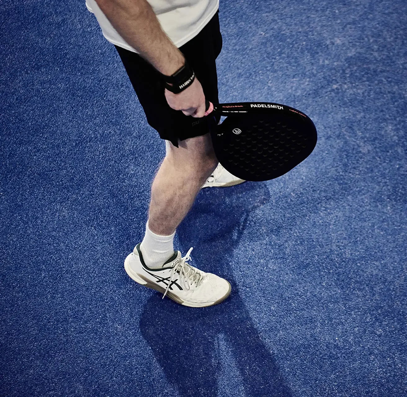 Person holding black padel racket on blue court, wearing sportswear and white sneakers