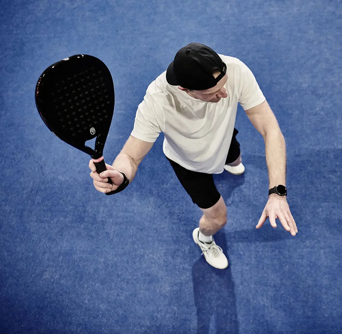 Man playing padel on blue court holding black carbon padel racket, wearing sportswear
